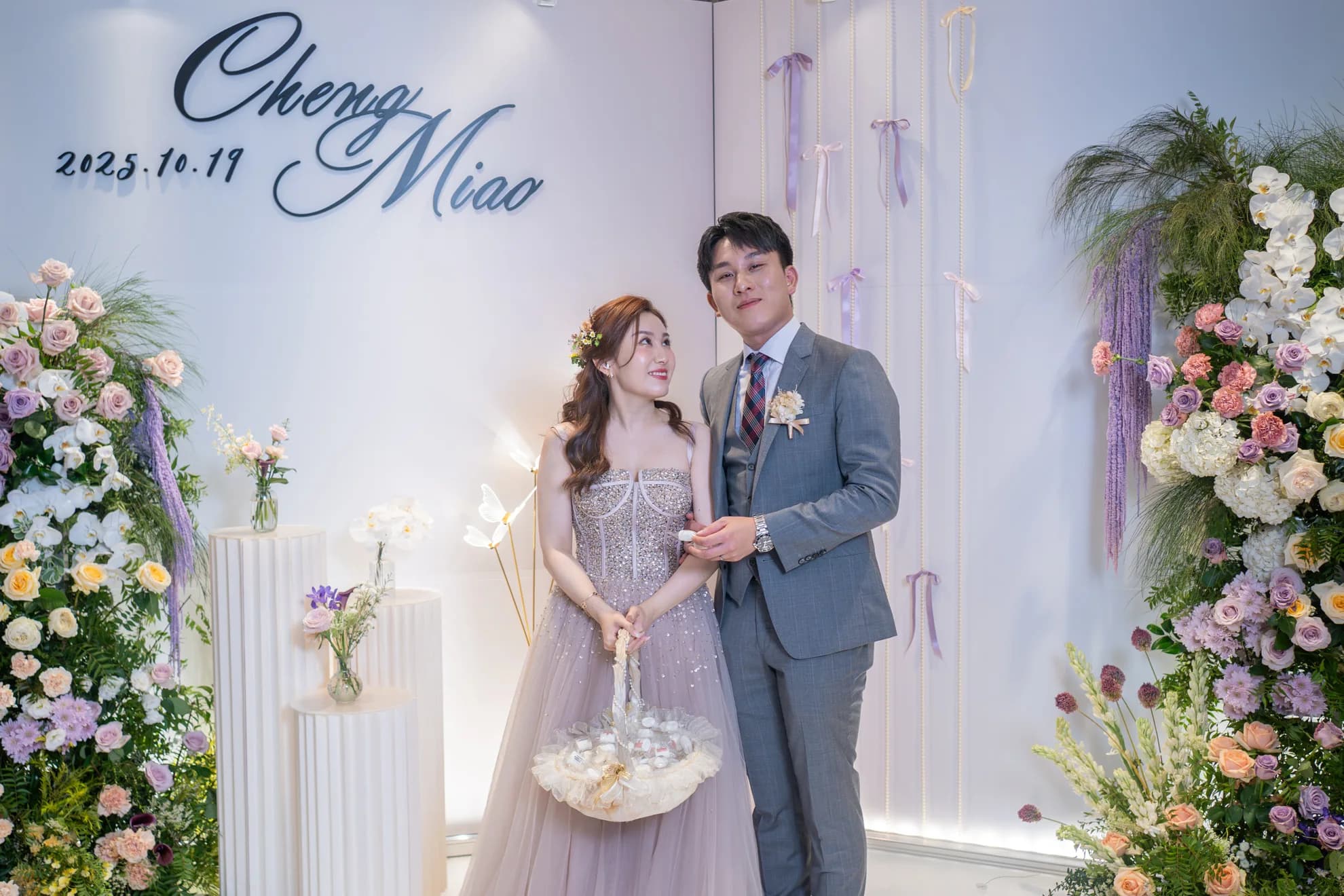 新婚夫婦於淺色花卉佈置前合影，牆上婚禮日期，整體色調溫馨浪漫。