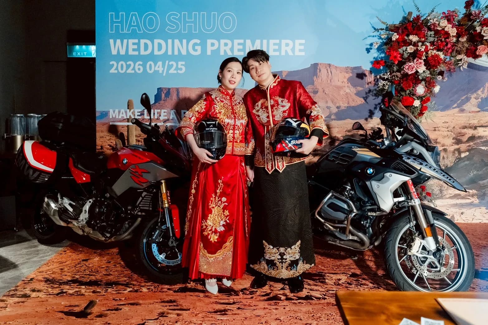 身著紅金中式禮服的新婚夫婦，手持頭盔與摩托車在沙漠背景前合影。