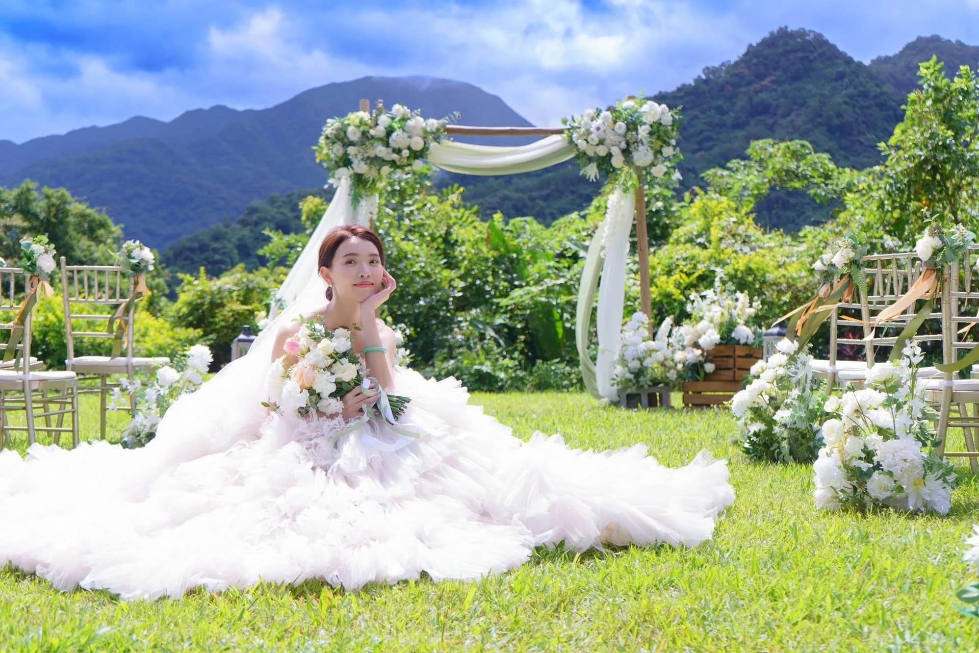 新娘坐於綠草地,手捧花束,白色婚紗與花藝拱門營造清新婚禮氛圍。