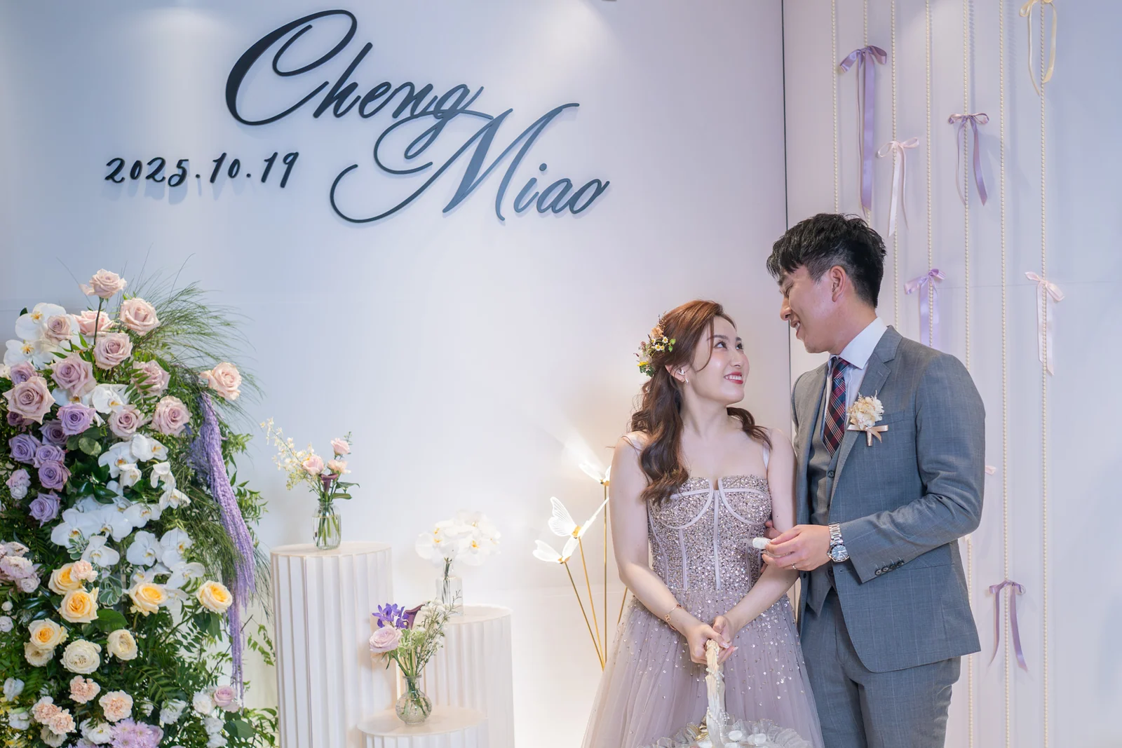 新人站在奶油白鮮花蝴蝶結背板前相視,兩側花藝保留中央合照空間